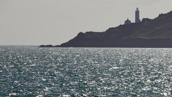 This landscape photograph depicts the Start Point Lighthouse situated prominently on the rugged coastline of Start Point in Devon, England, United Kingdom. Taken during the afternoon in early spring, the image features glistening sunlight reflecting off the water's surface in the foreground, while the lighthouse stands out against the silhouette of the coastal cliffs. The photograph captures the unique maritime atmosphere of the Devon coast with Start Point Lighthouse serving as a landmark of navigation and heritage.